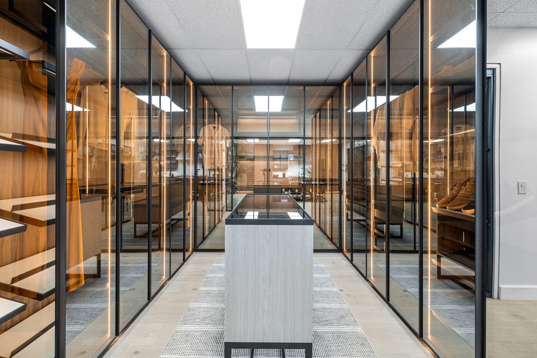 A long dressing-room corridor in glass-fronted timber cabinetry with a stone-topped island running its length, built by Fine Line.
