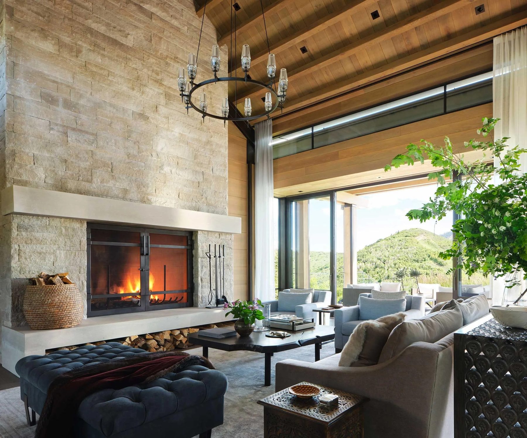A great room built by Fine Line: a full-height stone fireplace, timber ceiling and a glazed wall opening onto the hillside.